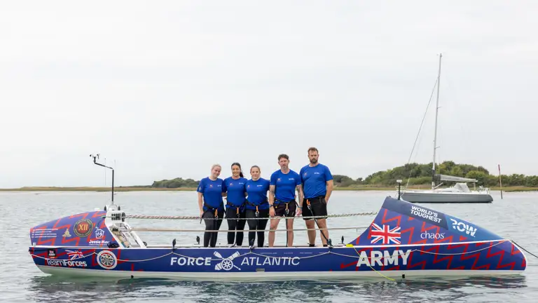 Five rowers in matching blue shirts stand on a sleek rowing boat labeled Force Atlantic Army on calm water.
