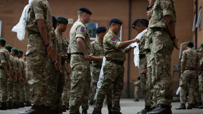 Groups of soldiers in camouflage uniforms stand in rows during a ceremony. Other soldiers are placing white scarves around some soldier's necks. The event is taking place outside near a brick building for a pass out parade.