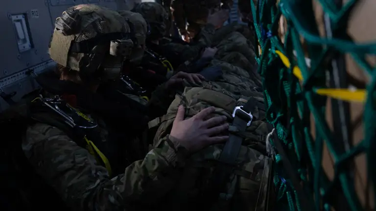 Soldiers wearing camouflage gear sit closely in a dimly lit aircraft, preparing for a jump. Netting on the right and teamwork convey focus and tension.