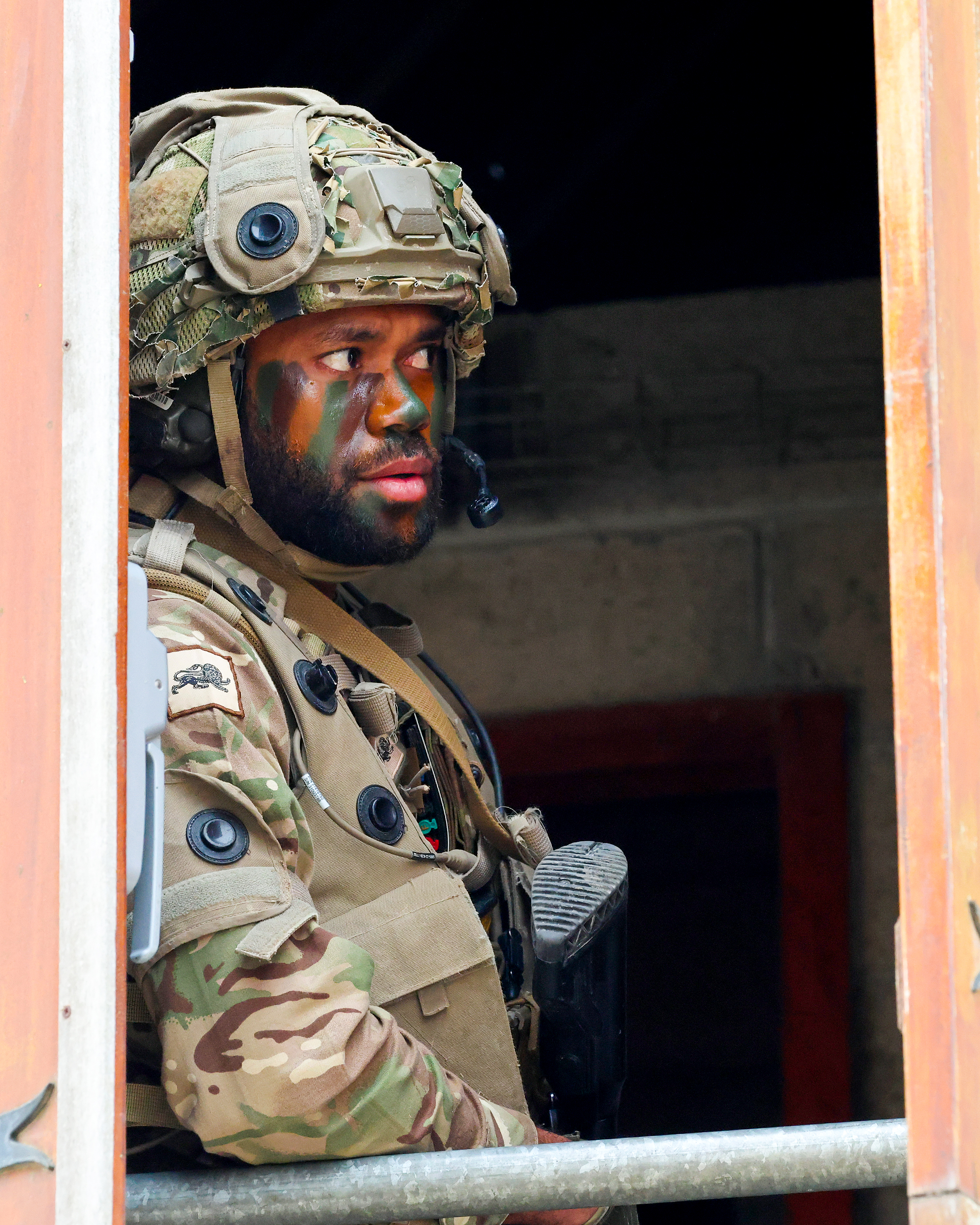 Soldier in full camouflage gear and helmet stands alert inside a building doorway, holding a rifle ready for action.
