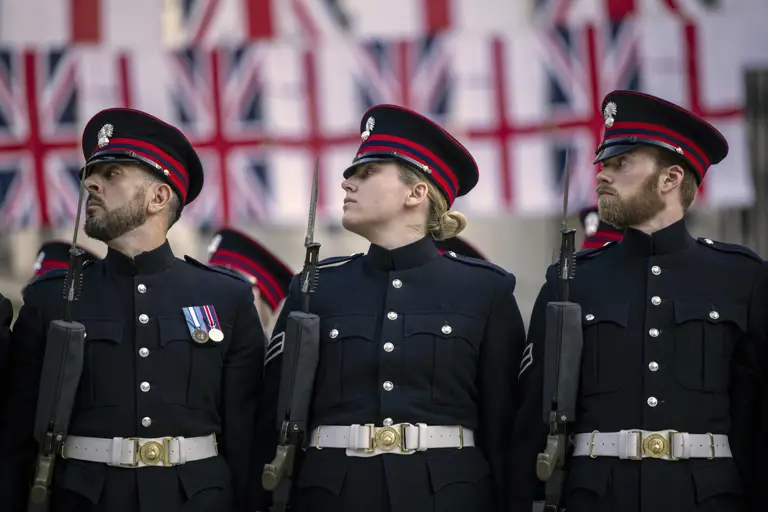 Three uniformed soldiers stand in formation holding rifles, with multiple Union Jack flags displayed in the background.