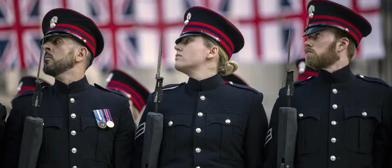Three uniformed soldiers stand in formation holding rifles, with multiple Union Jack flags displayed in the background.