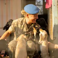 A female soldier in uniform and a blue beret is swarmed by several puppies.