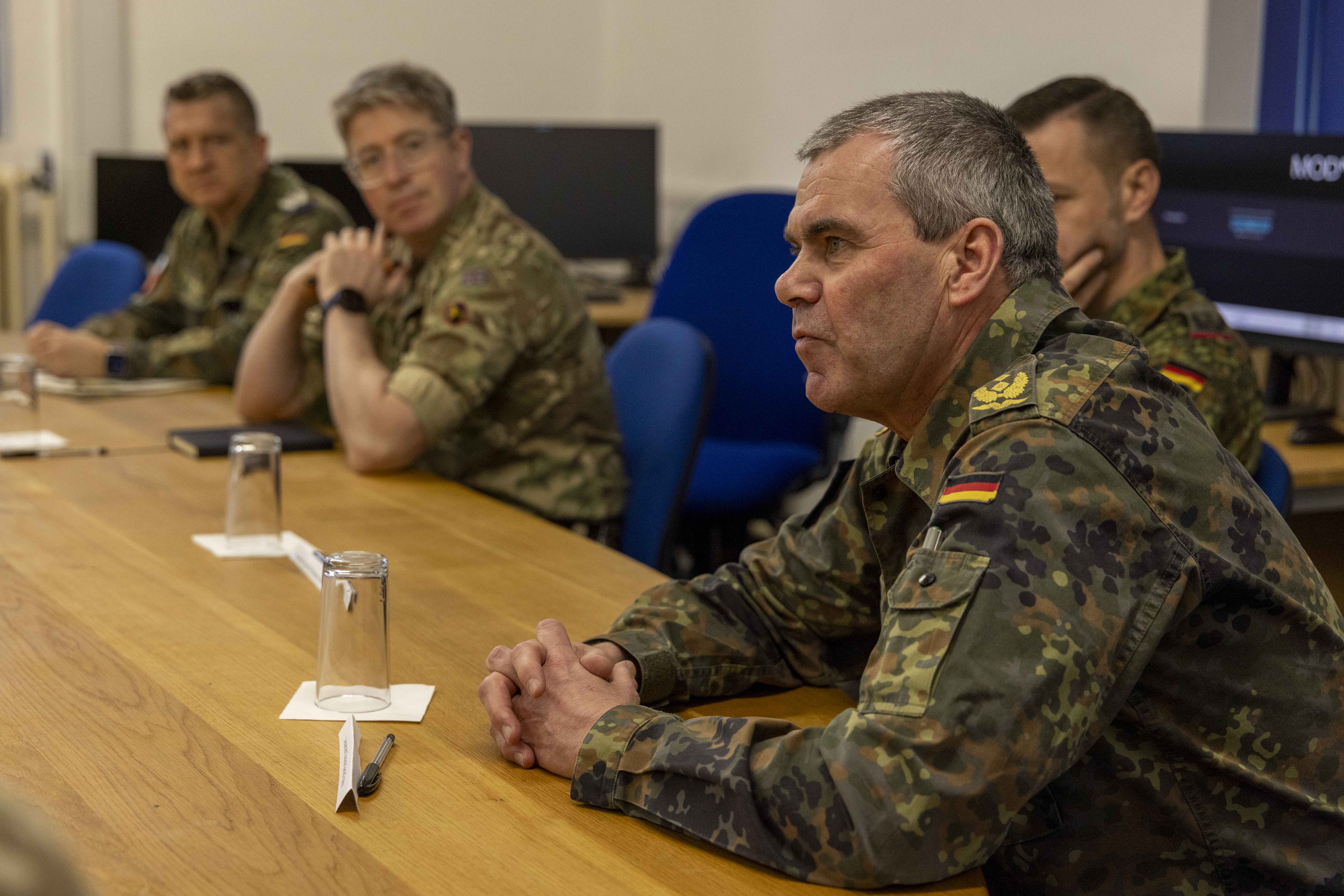A group of military personnel in camouflage uniforms sit attentively at a wooden conference table, engaged in discussion, with a serious atmosphere.