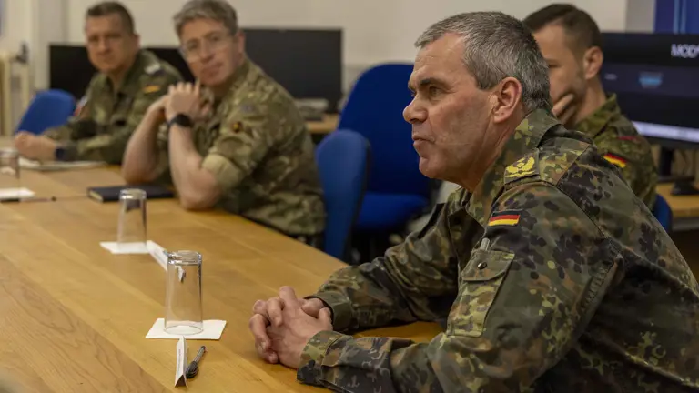 A group of military personnel in camouflage uniforms sit attentively at a wooden conference table, engaged in discussion, with a serious atmosphere.