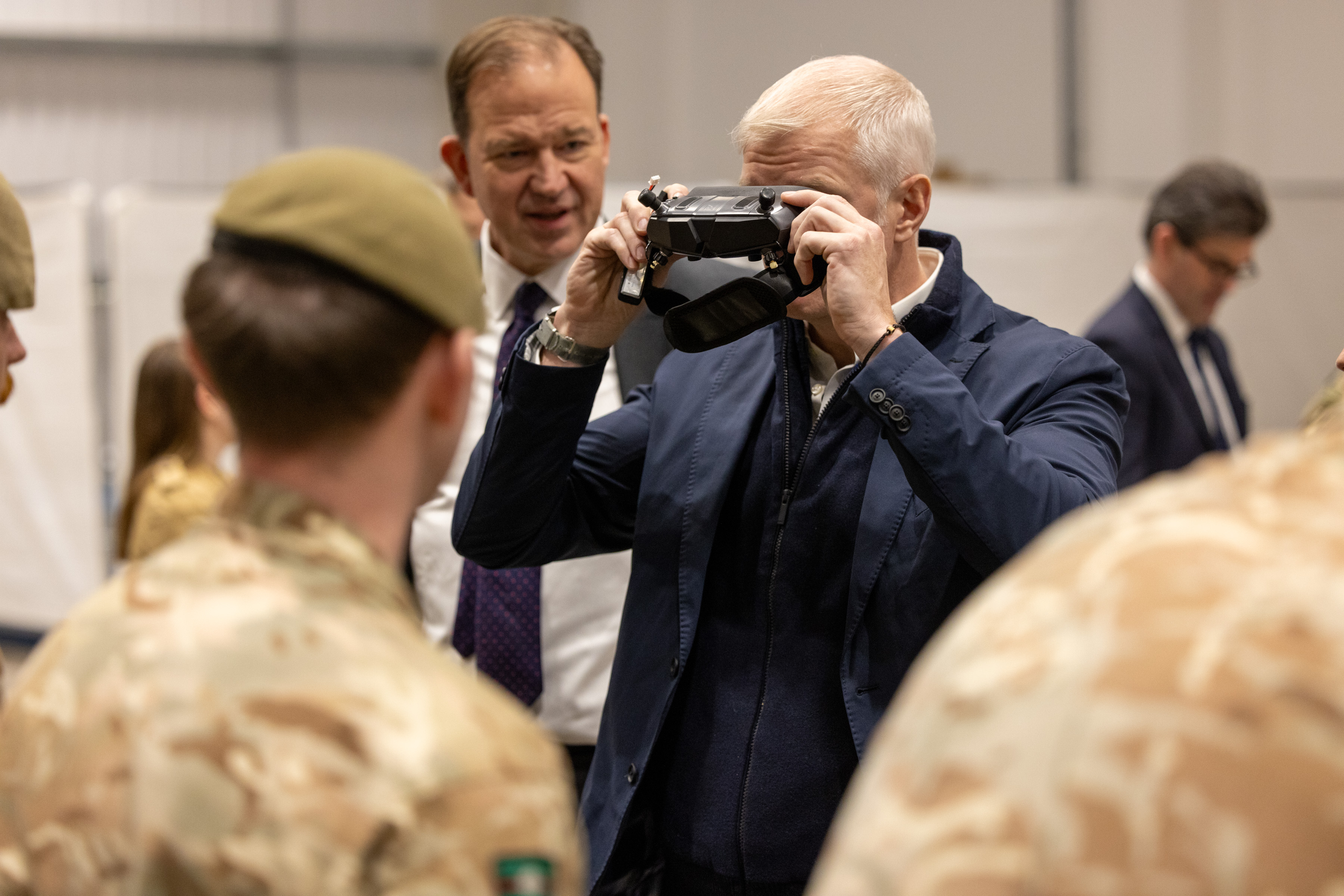 A man in a navy jacket uses a handheld device while surrounded by military personnel in camouflage uniforms indoors.