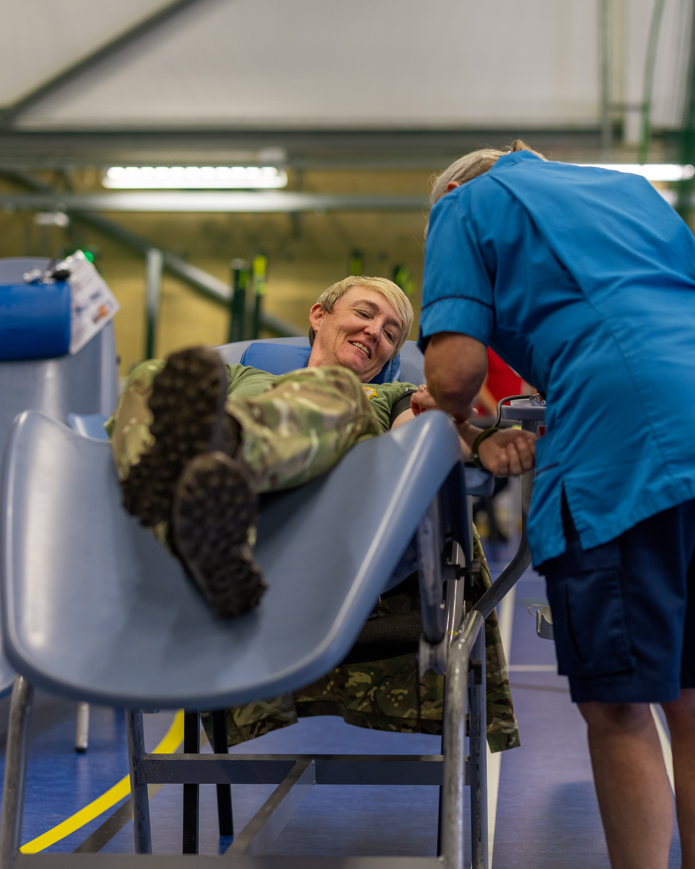A soldier in camouflage uniform reclines on a medical chair while a healthcare worker in blue scrubs attends to his arm.