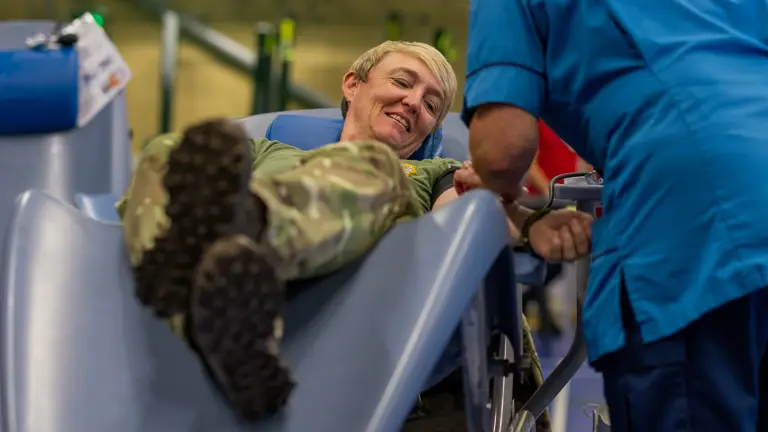 A soldier in camouflage uniform reclines on a medical chair while a healthcare worker in blue scrubs attends to his arm.