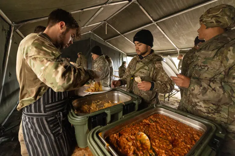 Soldiers in camouflage uniforms receiving hot meals served inside a large military tent during a field exercise.