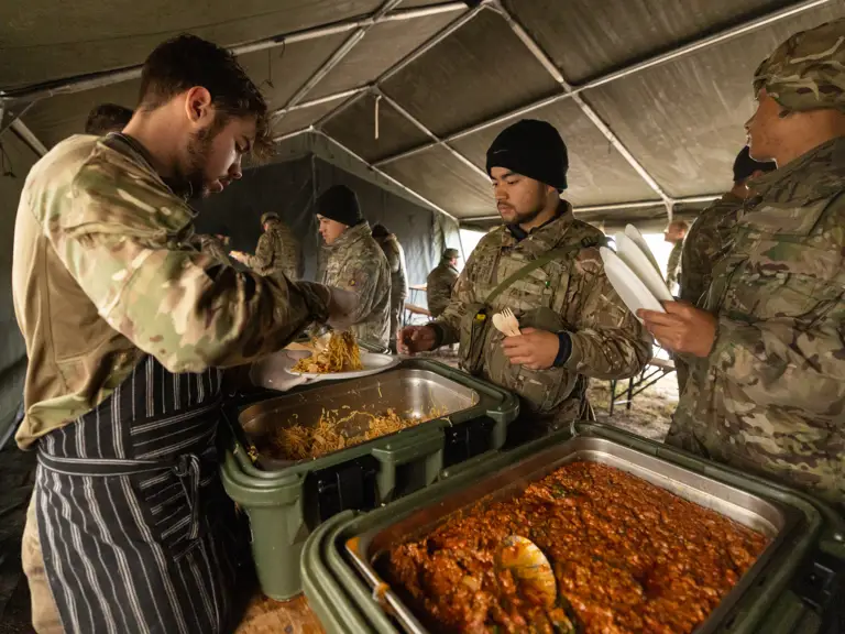 Soldiers in camouflage uniforms receiving hot meals served inside a large military tent during a field exercise.