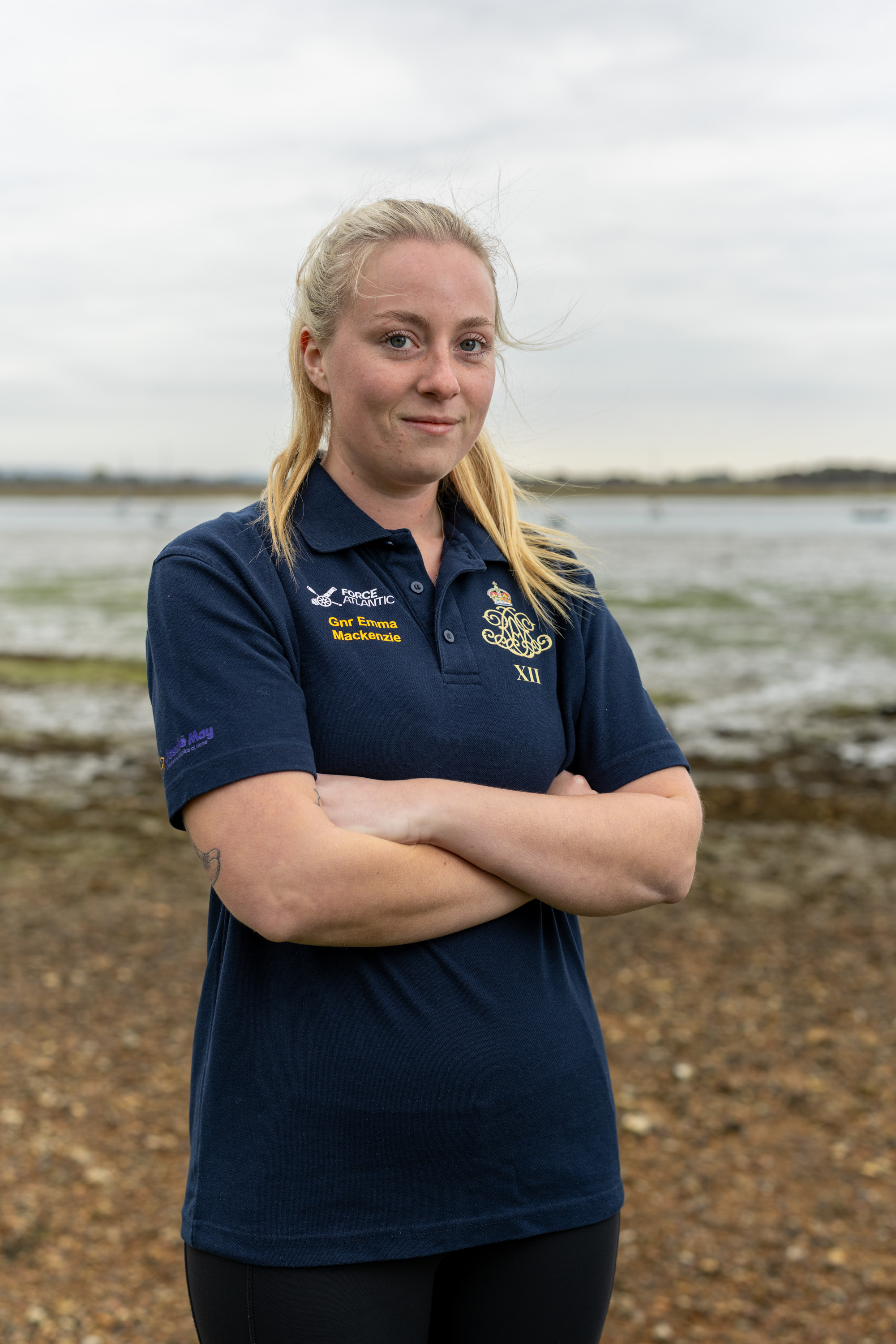 A female Army soldier, wearing dark blue shirt with an Army cap badge on it, is standing on a shoreline with an estuary in the background.