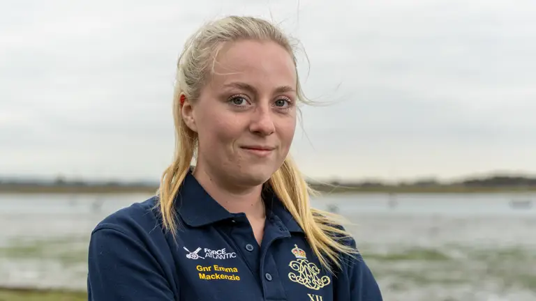 A female Army soldier, wearing dark blue shirt with an Army cap badge on it, is standing on a shoreline with an estuary in the background.