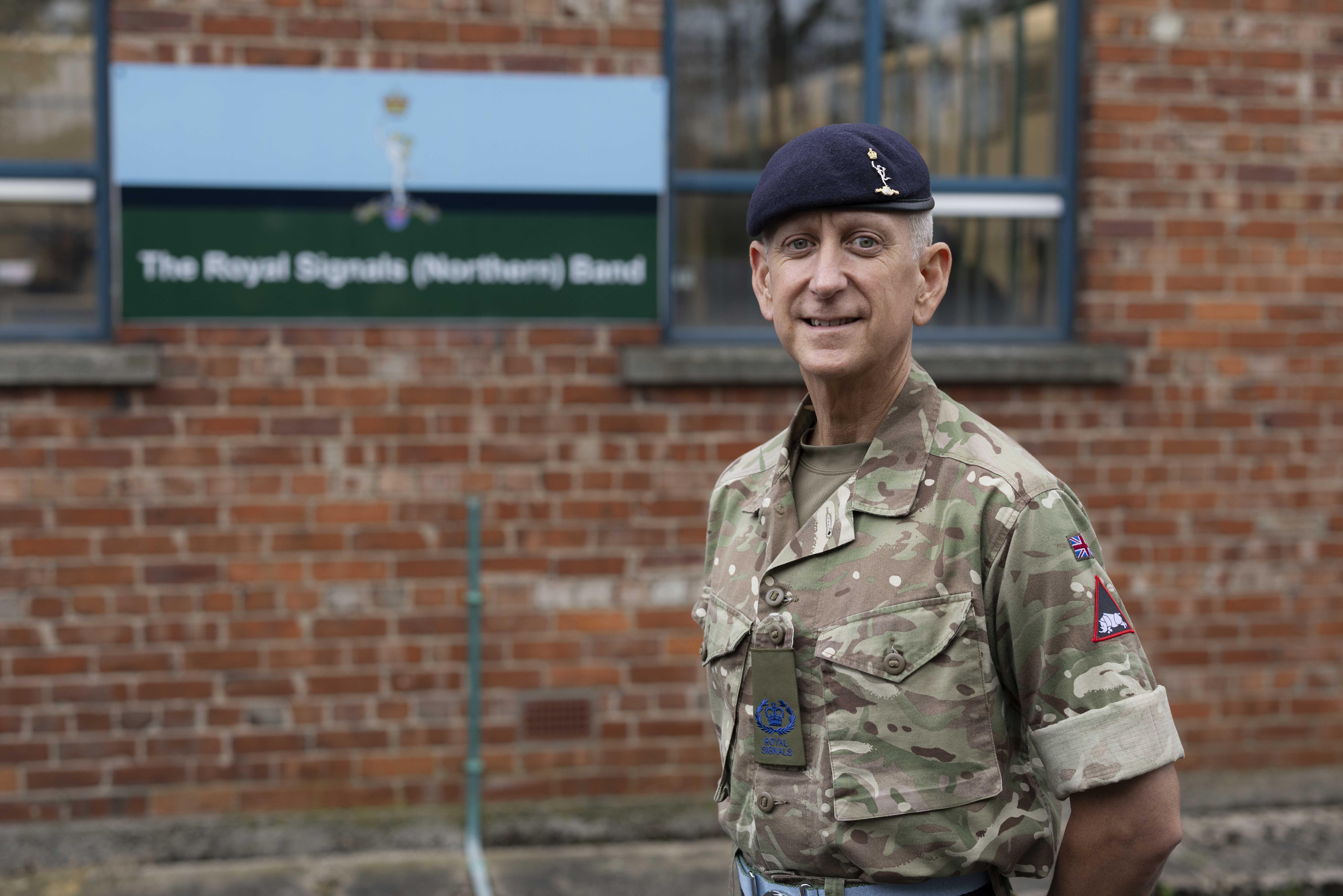 An individual in a military uniform stands in front of a sign for the Royal Signals (Northern) Band. The person is wearing a camouflage-patterned outfit and a dark beret.