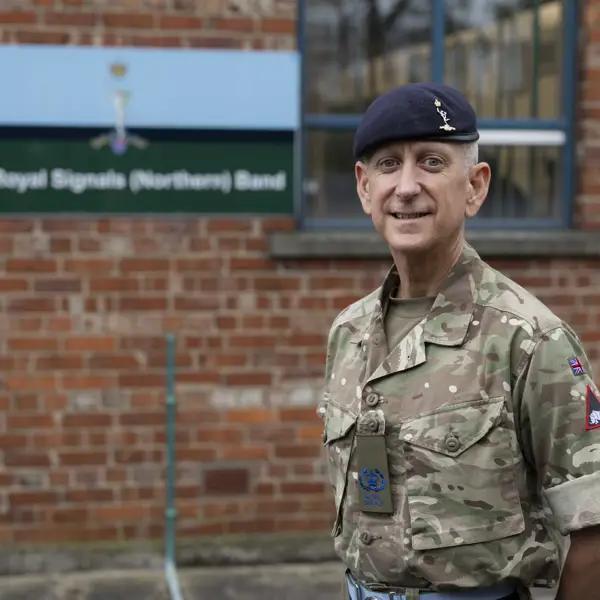 An individual in a military uniform stands in front of a sign for the Royal Signals (Northern) Band. The person is wearing a camouflage-patterned outfit and a dark beret.