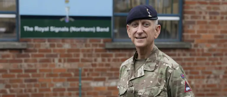 An individual in a military uniform stands in front of a sign for the Royal Signals (Northern) Band. The person is wearing a camouflage-patterned outfit and a dark beret.