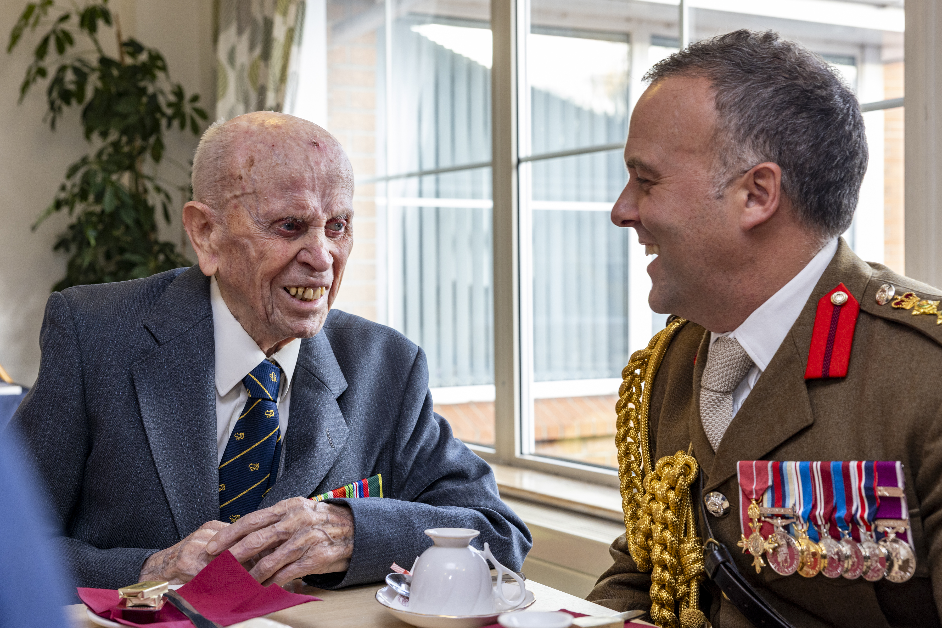 Two men in formal attire, one elderly veteran with medals and the other in a decorated military uniform, engaged in conversation at a table.