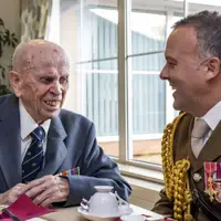 Two men in formal attire, one elderly veteran with medals and the other in a decorated military uniform, engaged in conversation at a table.