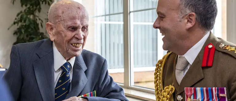 Two men in formal attire, one elderly veteran with medals and the other in a decorated military uniform, engaged in conversation at a table.