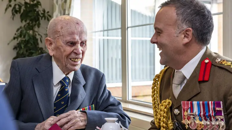 Two men in formal attire, one elderly veteran with medals and the other in a decorated military uniform, engaged in conversation at a table.