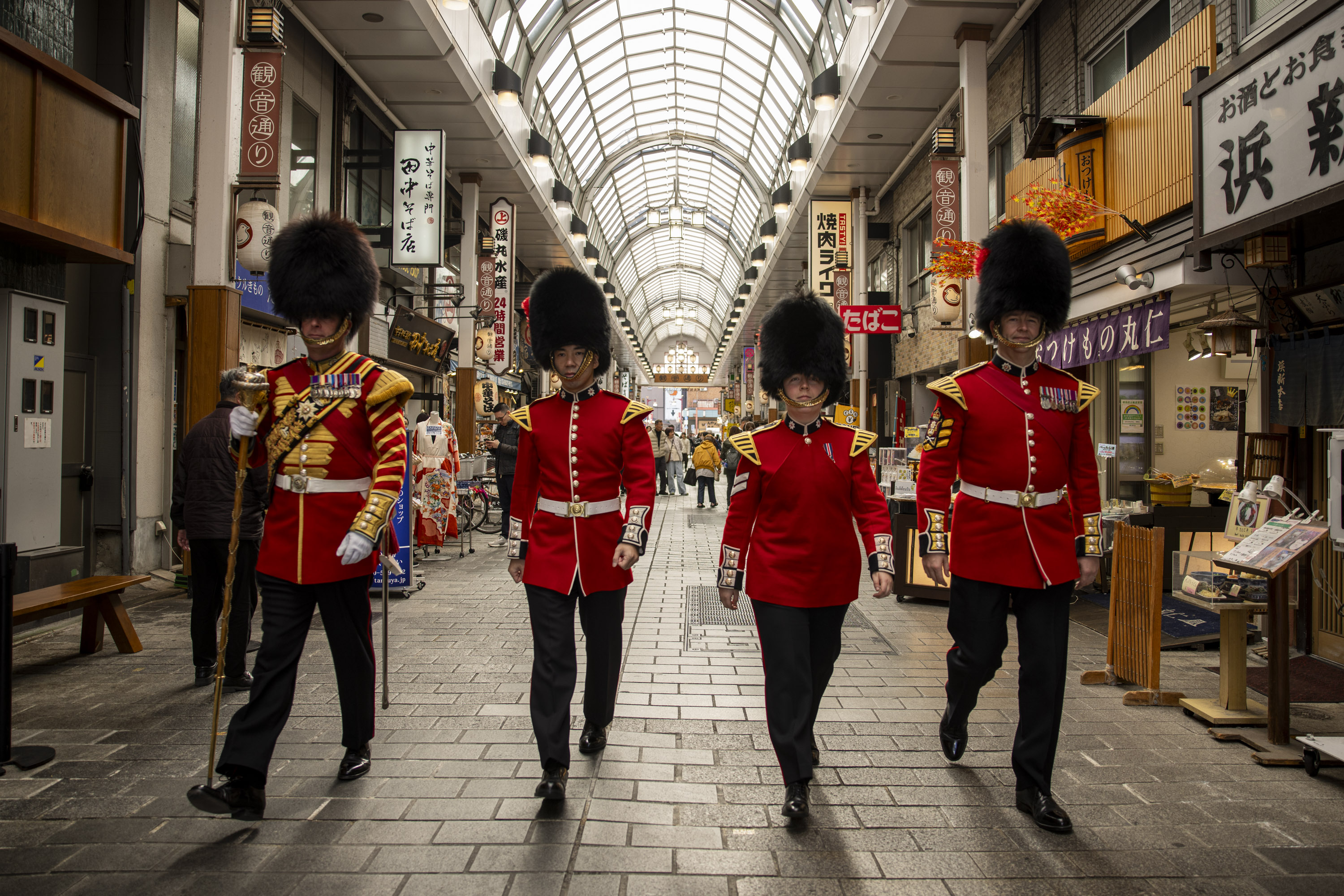 Four soldiers in red tunics and bearskin hats walk through a Japanese shopping centre. 