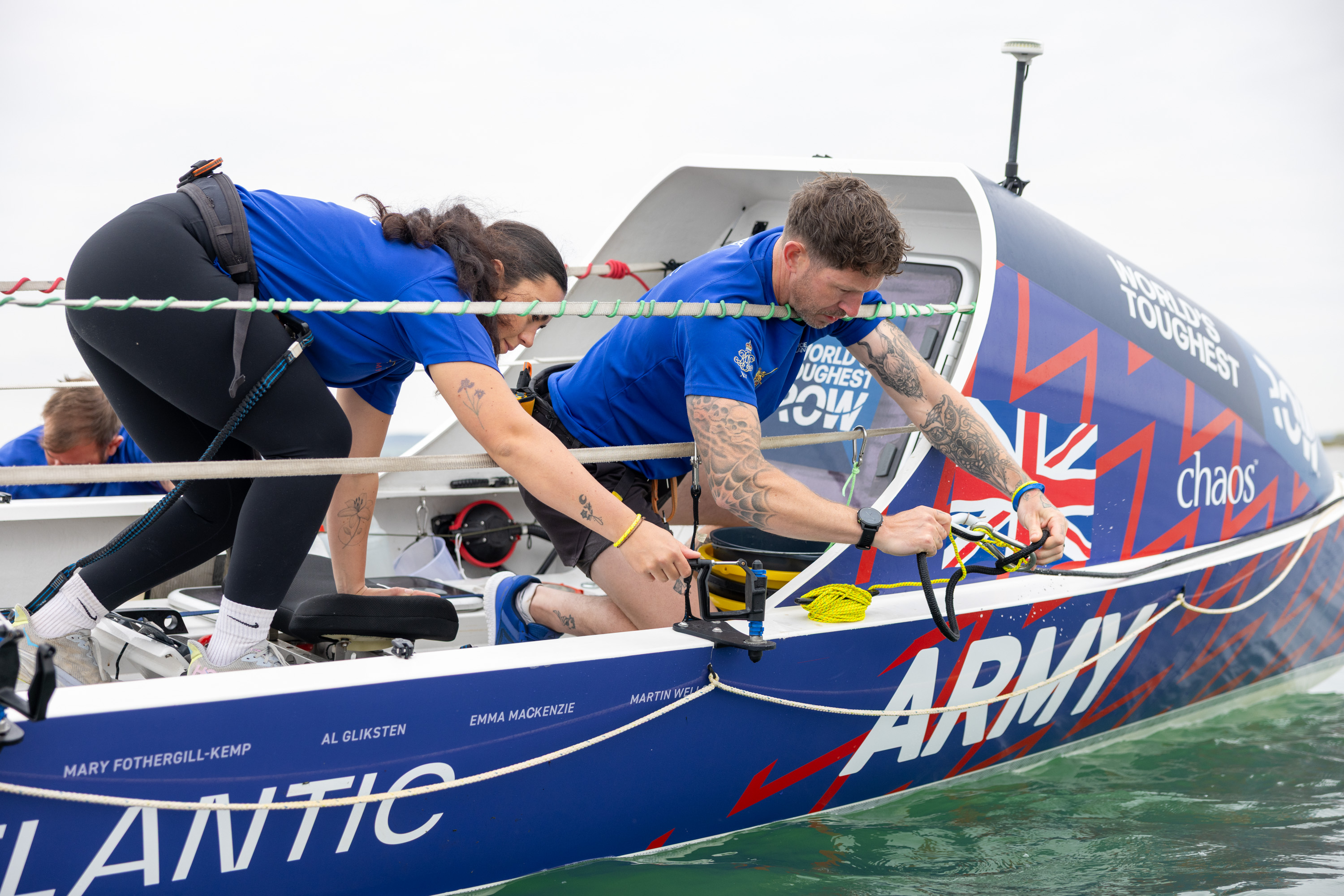 Two rowers in blue uniforms prepare their boat labeled 'Army' and 'Atlantic' on calm water.