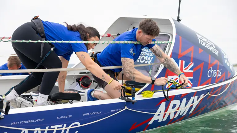 Two rowers in blue uniforms prepare their boat labeled 'Army' and 'Atlantic' on calm water.