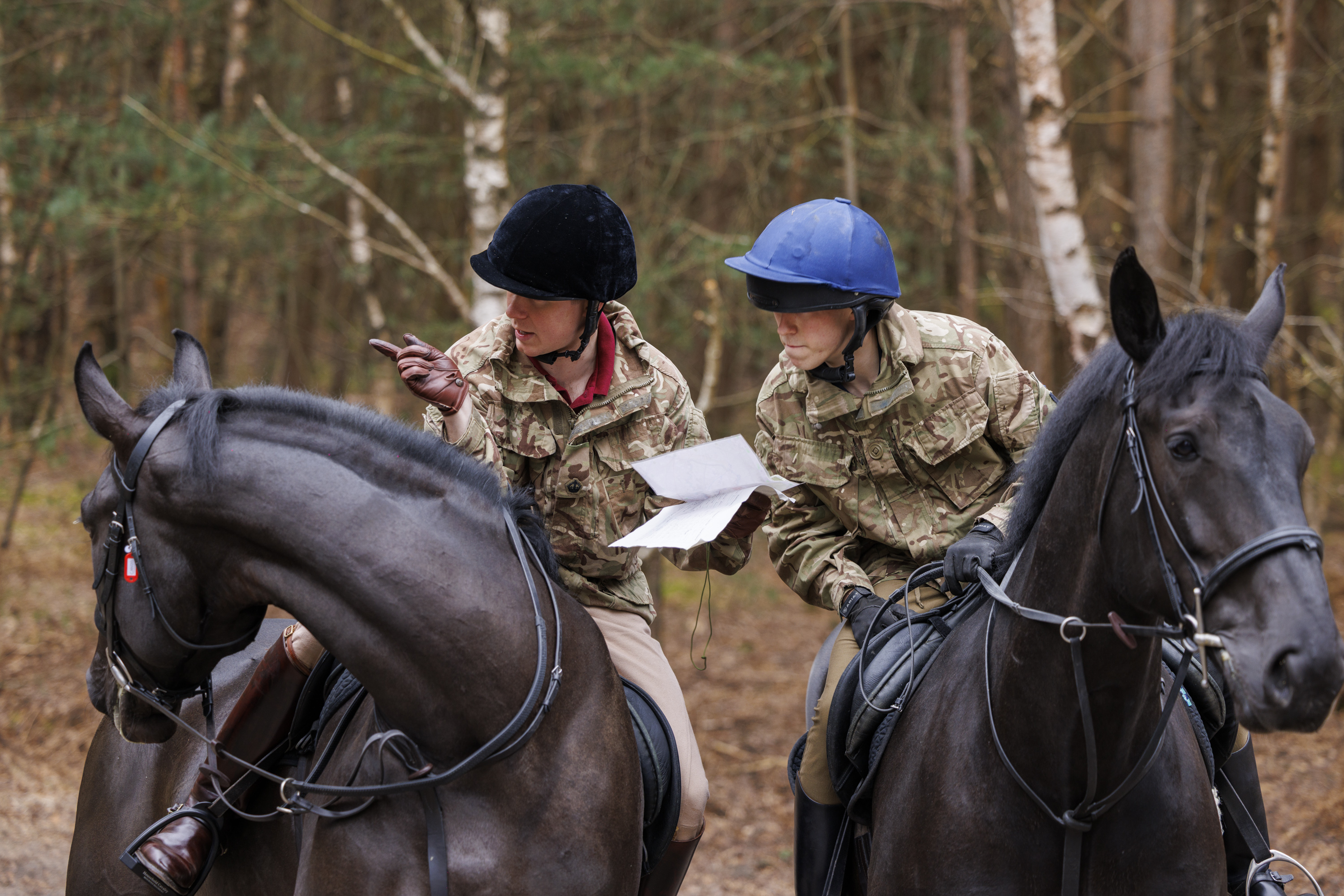 Two people in camouflage uniforms are riding black horses through a forested area. They are both wearing helmets.