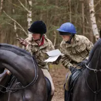 Two people in camouflage uniforms are riding black horses through a forested area. They are both wearing helmets.