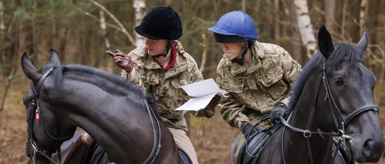 Two people in camouflage uniforms are riding black horses through a forested area. They are both wearing helmets.