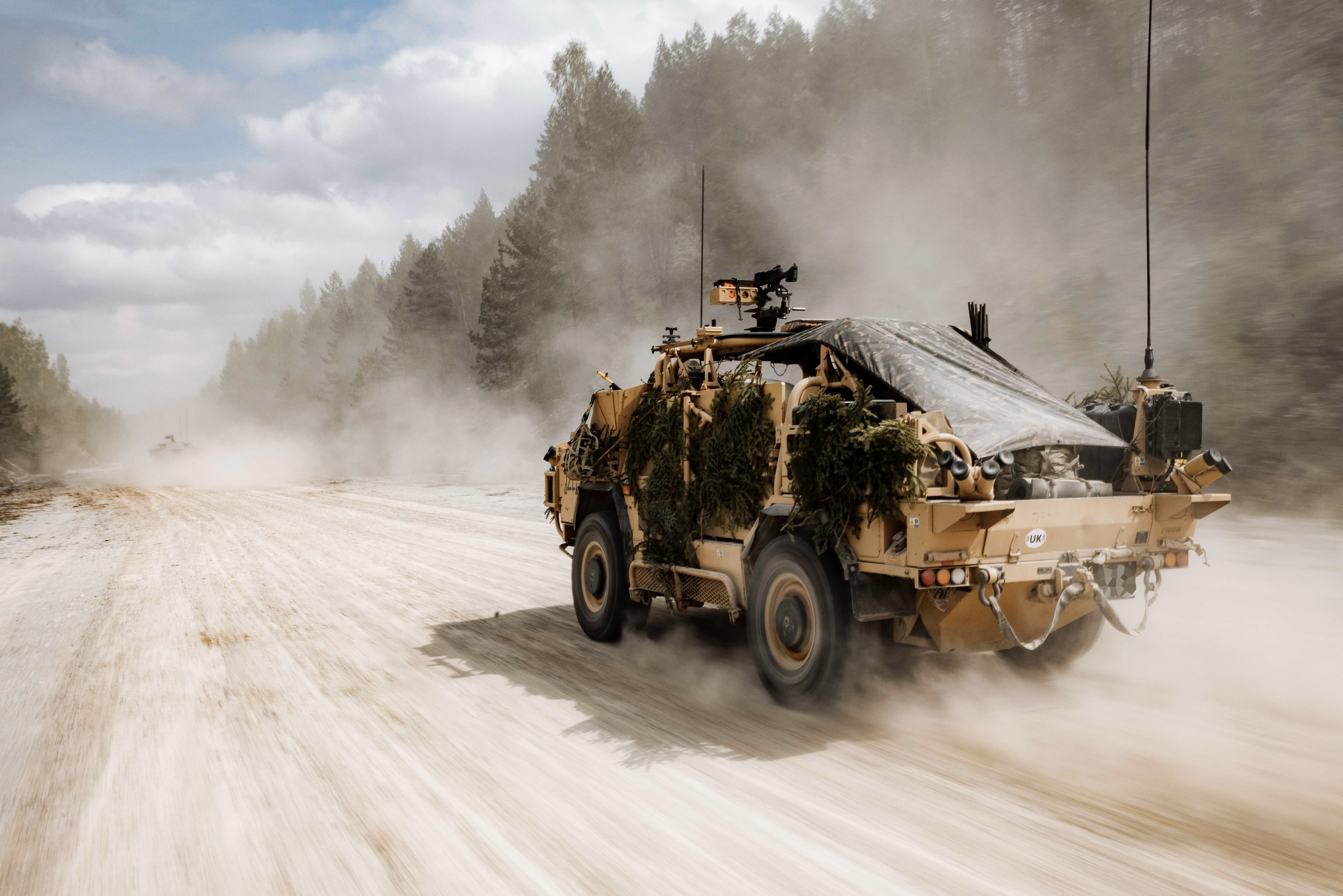 An armored military vehicle covered with foliage is driving on a sandy road surrounded by trees, creating a dusty trail.