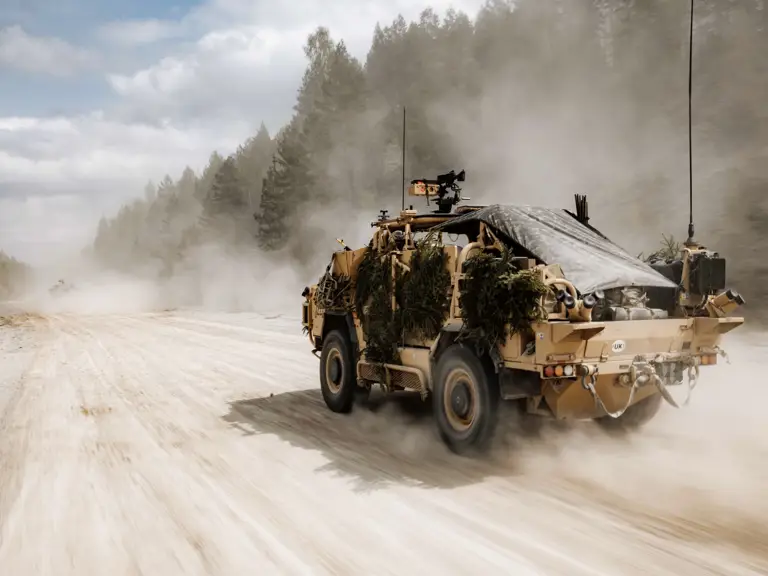 An armored military vehicle covered with foliage is driving on a sandy road surrounded by trees, creating a dusty trail.