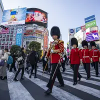 Four soldiers in red tunics and bearskin hats are pictured walking across a crossing in Tokyo.