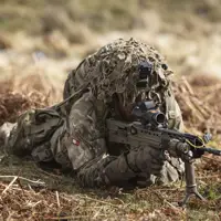 Soldier is seen wearing camouflage uniform including helmet and body armour, laying on his stomach in a training area, looking through the scoop of his rifle on exercise
