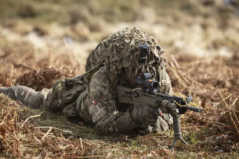 Soldier is seen wearing camouflage uniform including helmet and body armour, laying on his stomach in a training area, looking through the scoop of his rifle on exercise