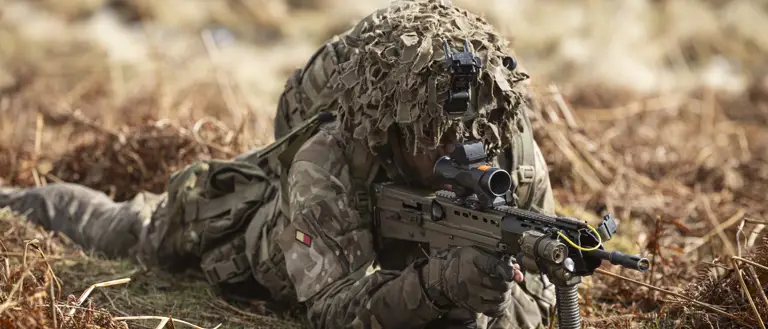 Soldier is seen wearing camouflage uniform including helmet and body armour, laying on his stomach in a training area, looking through the scoop of his rifle on exercise