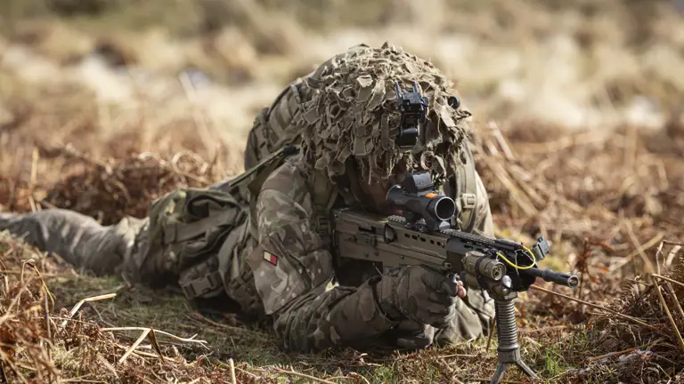 Soldier is seen wearing camouflage uniform including helmet and body armour, laying on his stomach in a training area, looking through the scoop of his rifle on exercise