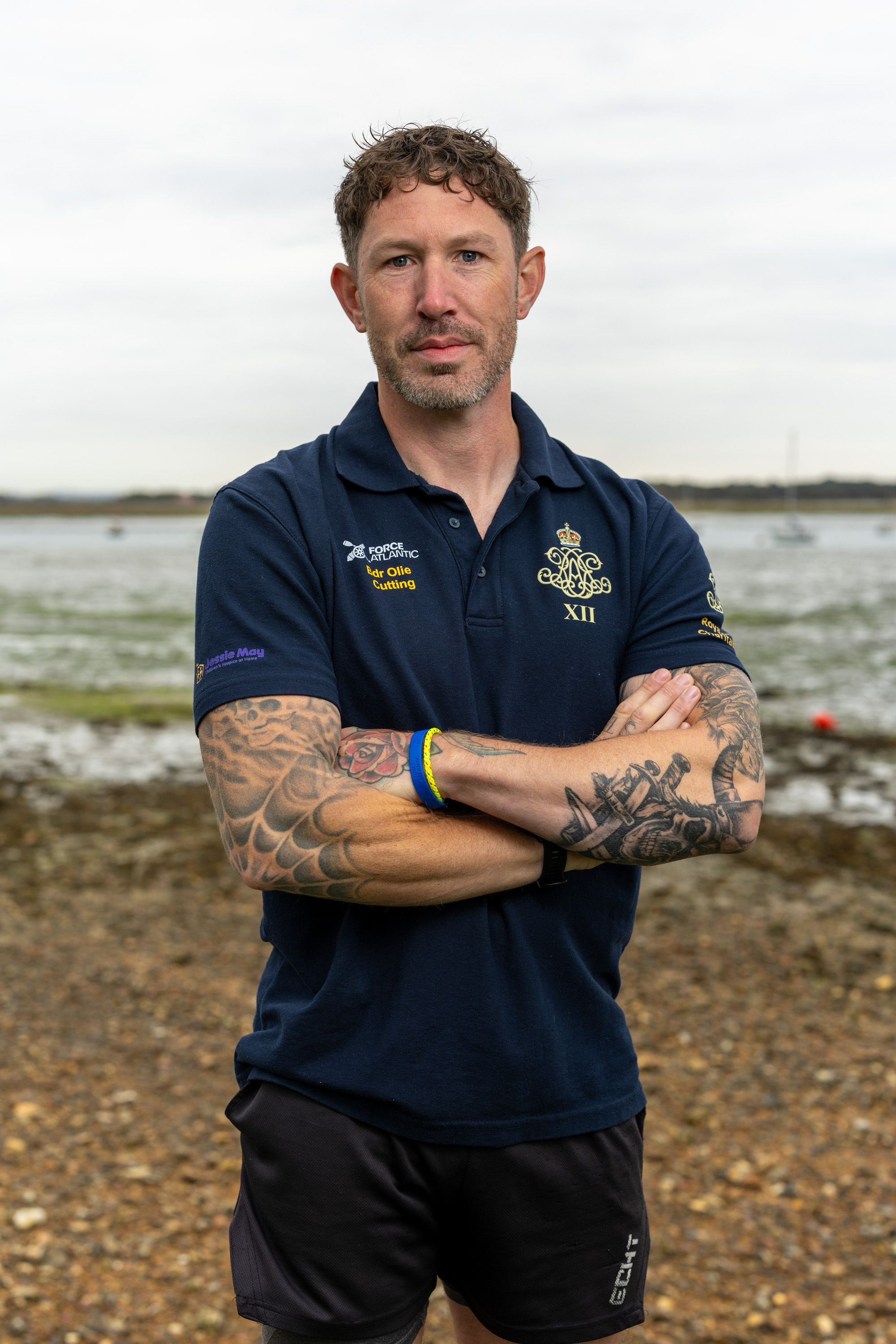 Man with tattooed arms wearing a navy polo shirt, stands with arms crossed on a rocky shore.