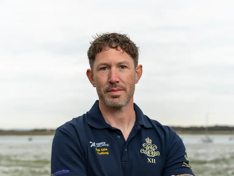 Man with tattooed arms wearing a navy polo shirt, stands with arms crossed on a rocky shore.
