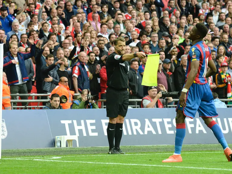 A football match scene showing an official on the side of the pitch holding a yellow flag.