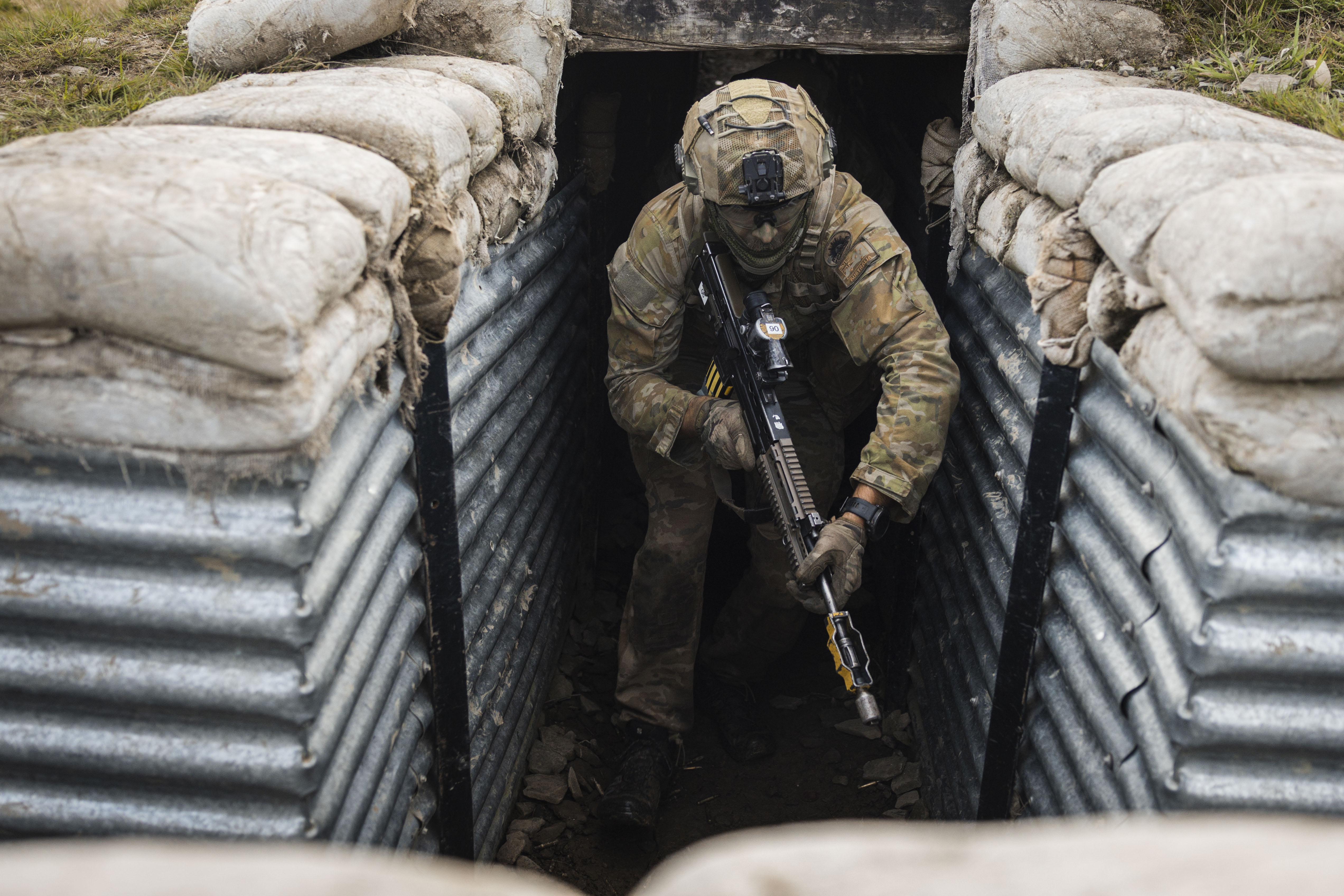 A soldier in camouflage gear crouches with a rifle while exiting a sandbag-lined trench.
