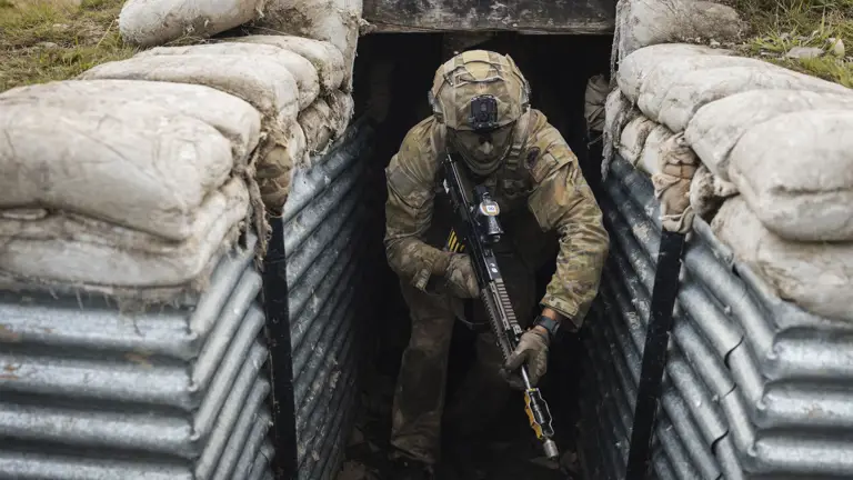A soldier in camouflage gear crouches with a rifle while exiting a sandbag-lined trench.