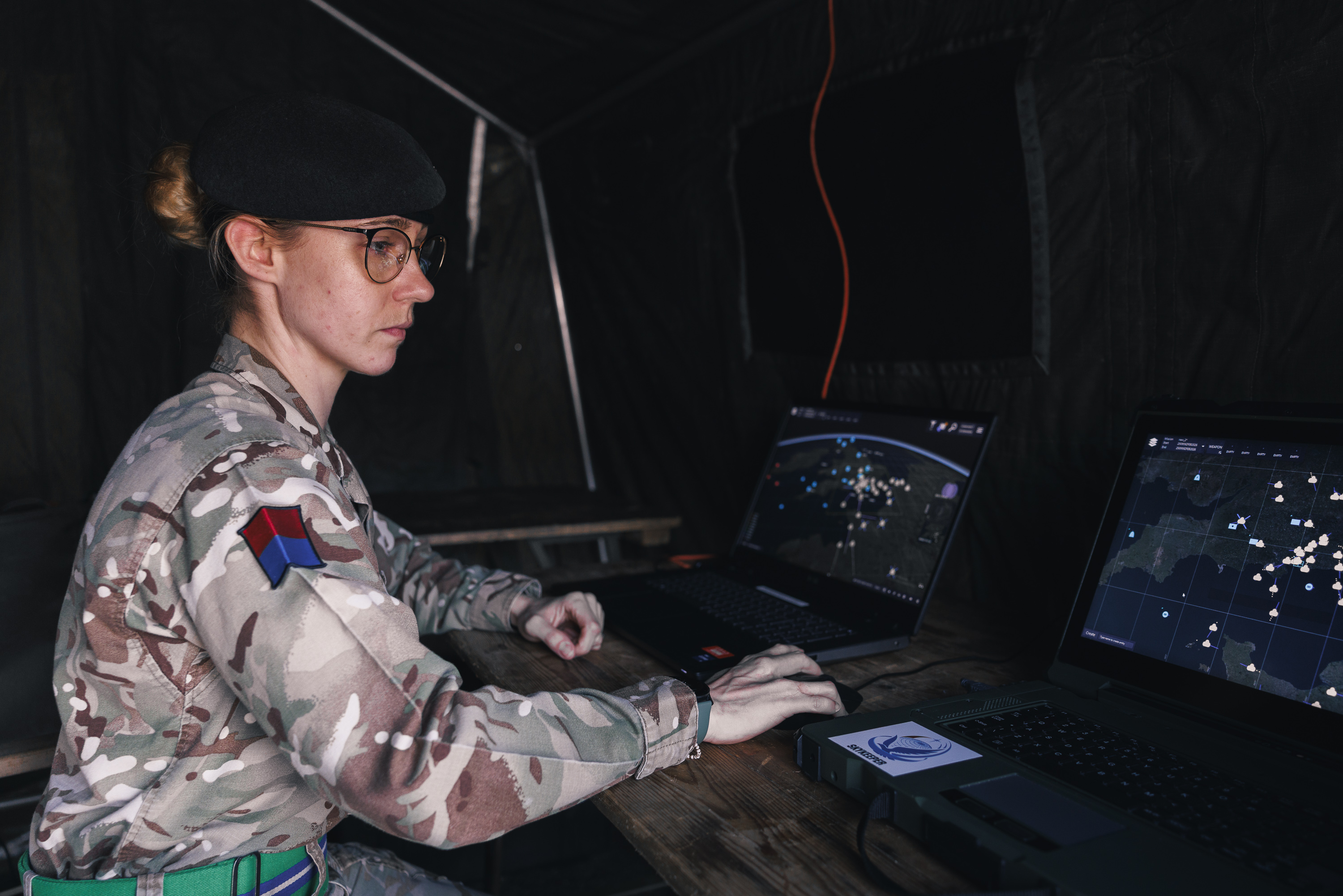 A person in military camouflage attire works on two laptops inside a dark tent. The screens display maps with various data points. A red and blue patch is visible on the uniform.