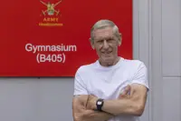 A man wearing a white t-shirt stands in front of a red sign which reads 'Army Headquarters'.