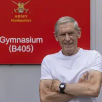 A man wearing a white t-shirt stands in front of a red sign which reads 'Army Headquarters'.