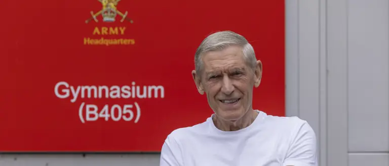 A man wearing a white t-shirt stands in front of a red sign which reads 'Army Headquarters'.