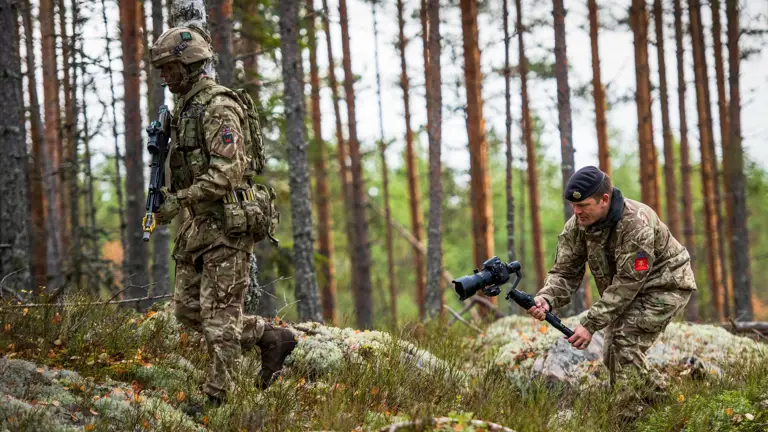 An Army Photographer films a British Army infantry soldier walking through the woods. The infanteer is wearing body armour, helmet and webbing while carrying his SA80 rifles. The Army Photographer is filming on an Nikon which is mounted to a DJI Ronin gimbal.