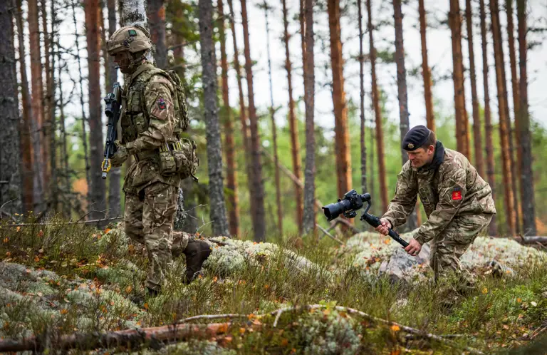 An Army Photographer films a British Army infantry soldier walking through the woods. The infanteer is wearing body armour, helmet and webbing while carrying his SA80 rifles. The Army Photographer is filming on an Nikon which is mounted to a DJI Ronin gimbal.