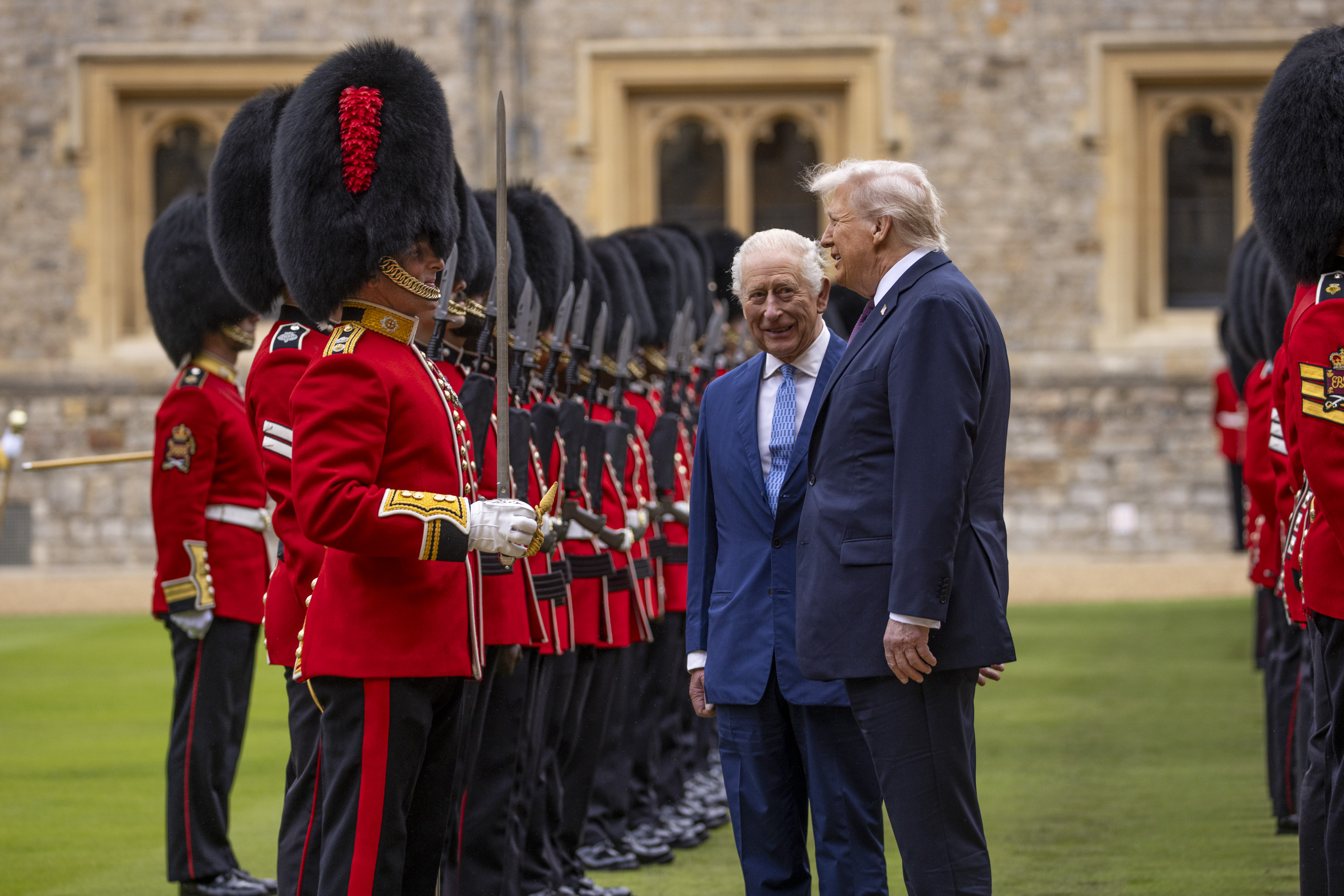 Two men in suits inspect a line of British guards dressed in traditional red uniforms and bearskin hats.