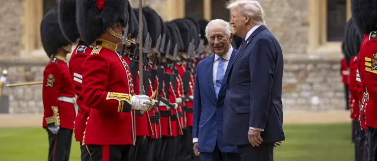 Two men in suits inspect a line of British guards dressed in traditional red uniforms and bearskin hats.