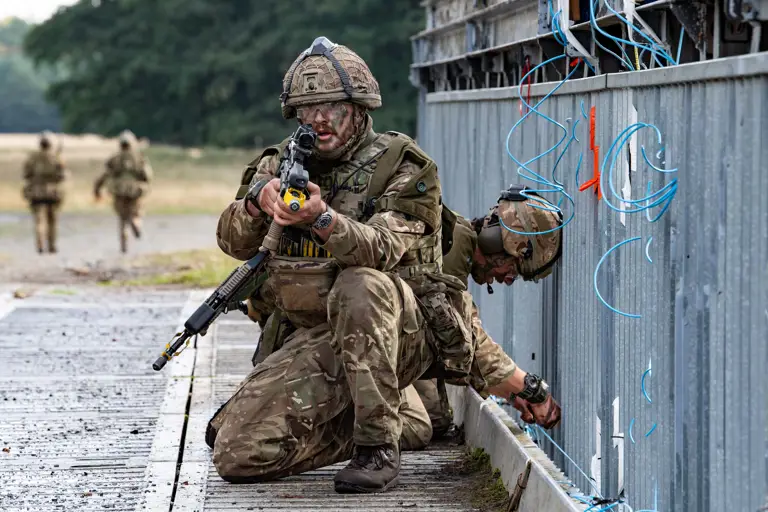 Two soldiers in camouflage gear conducting a tactical operation near a metal structure with exposed wiring outdoors.